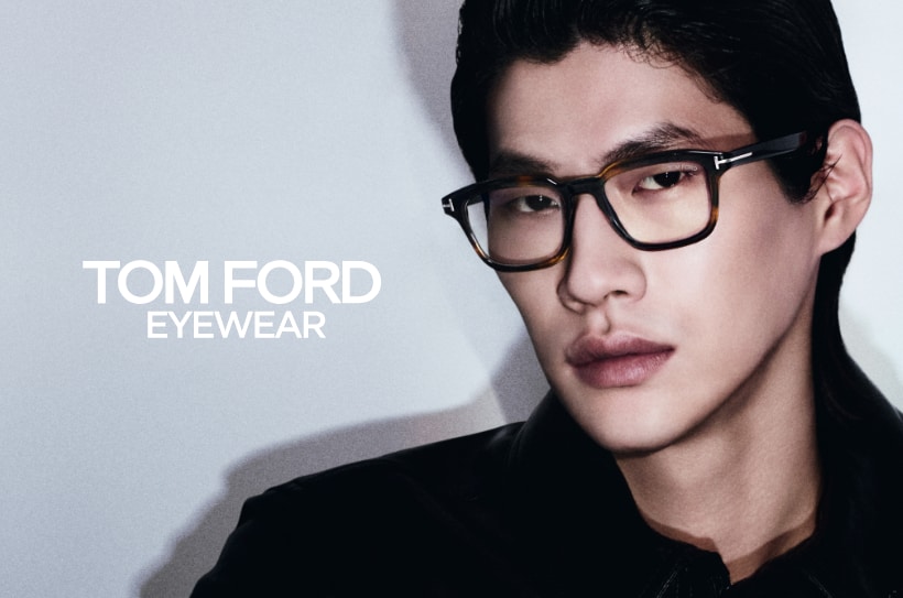 Man met een havanna-bril, staand naast de tekst \'Tom Ford Eyewear\' op een lichte achtergrond