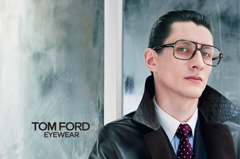 Homme portant des lunettes Tom Ford, vêtu d’un manteau en cuir et d’une cravate, devant un fond moderne.