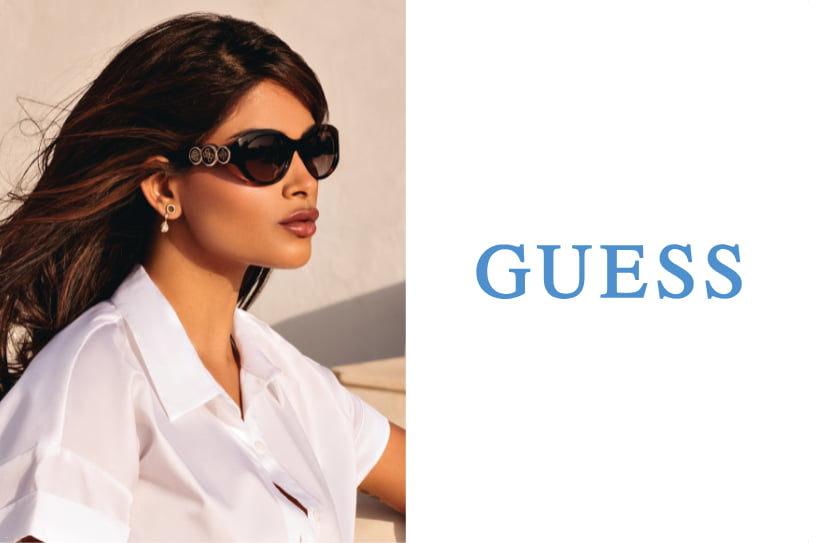 Femme portant des lunettes de soleil Guess noires et un chemisier blanc dans un décor ensoleillé.