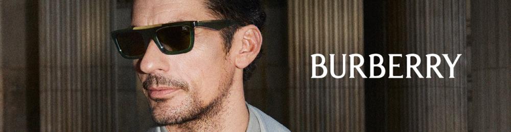 Homme portant des lunettes de soleil Burberry foncées et un manteau gris clair, devant une façade à colonnes; logo Burberry au centre de l’image.