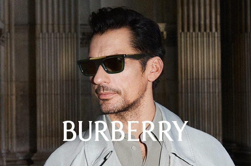 Homme portant des lunettes de soleil Burberry foncées et un manteau gris clair, devant une façade à colonnes; logo Burberry au centre de l’image.