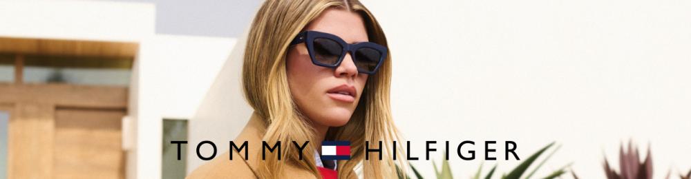 Vrouw met blond haar draagt een zwarte zonnebril. Logo Tommy Hilfiger zichtbaar.