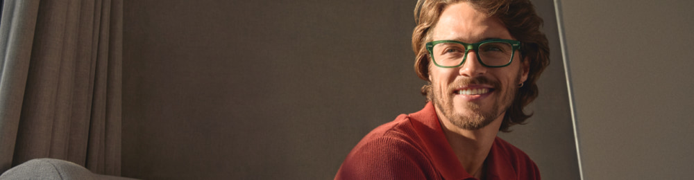 Lachende man met een groene bril en rood poloshirt zit ontspannen op een bank.