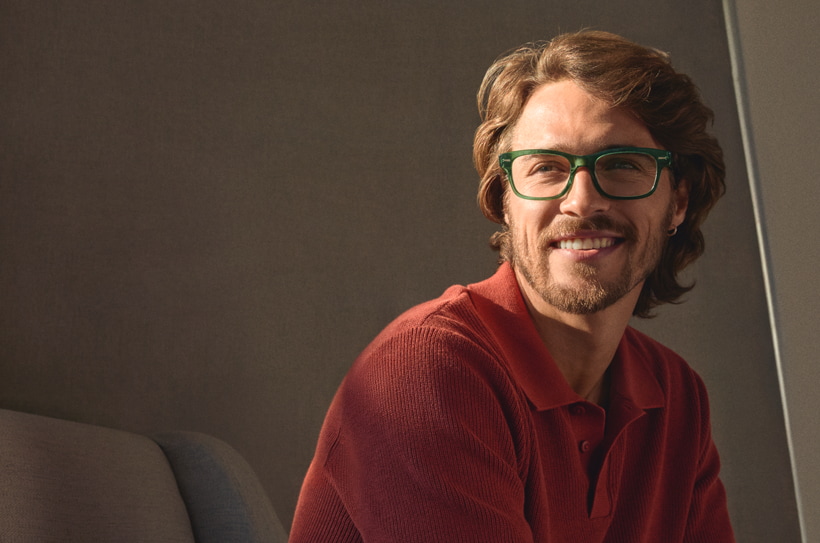 Lachende man met een groene bril en rood poloshirt zit ontspannen op een bank.