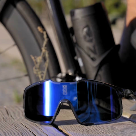 Lunettes de soleil Chamelo avec verres bleus réfléchissants, posées sur une surface en bois à côté d’un vélo.
