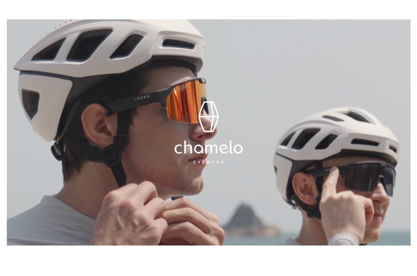 Twee fietsers met witte helmen dragen Chamelo zonnebrillen, klaar voor een rit, met Chamelo-logo in het midden.