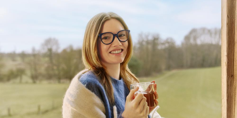 Vrouw met blauwe bril lacht en houdt een kop thee vast, buiten met groen landschap op de achtergrond.