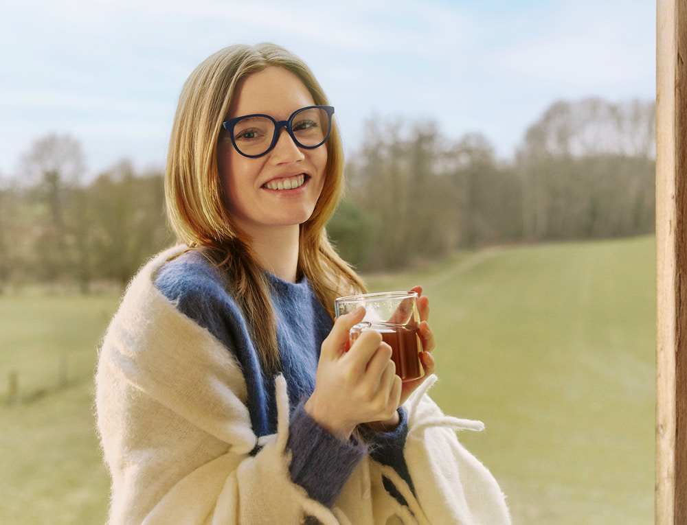 Vrouw met blauwe bril lacht en houdt een kop thee vast, buiten met groen landschap op de achtergrond.