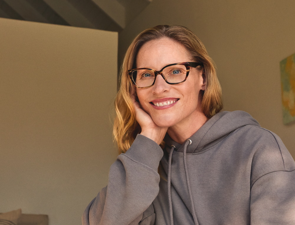 Een glimlachende vrouw met een bril en een grijze hoodie, zittend in een goed verlichte kamer met abstracte schilderijen.