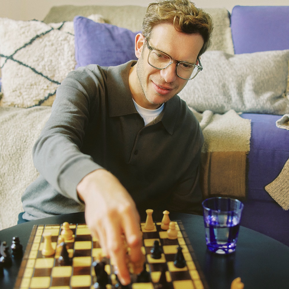 Man met bril speelt schaak in een woonkamer, ontspannen sfeer met paarse kussens op de achtergrond.