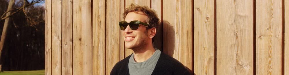 Man met zonnebril leunt ontspannen tegen een houten wand, lachend in de zon met groen grasveld op de achtergrond.