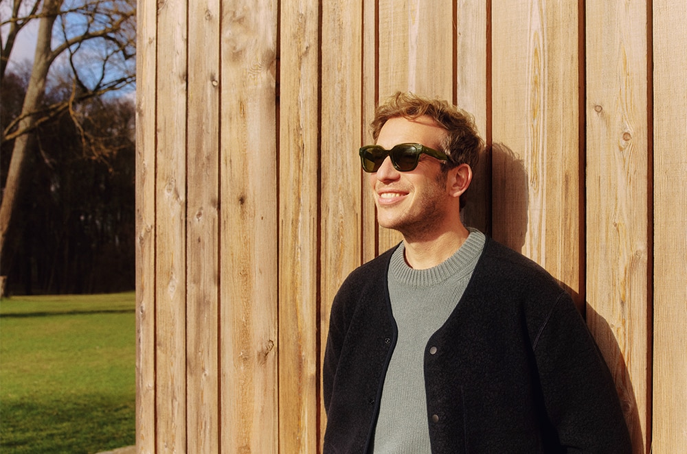 Man met zonnebril leunt ontspannen tegen een houten wand, lachend in de zon met groen grasveld op de achtergrond.