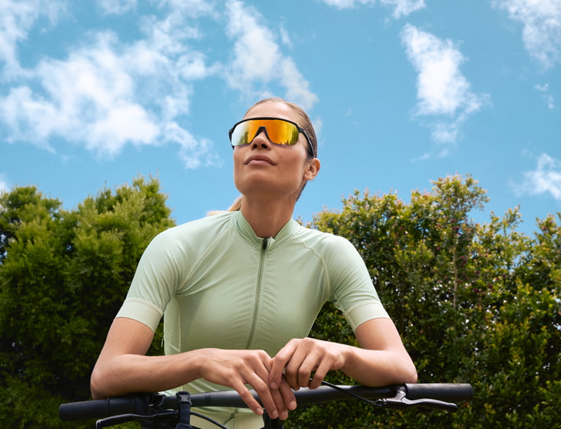 Een sportieve vrouw in zonnebrillen en groen fietsoutfit, grip houdend op haar fiets tegen een groene achtergrond.
