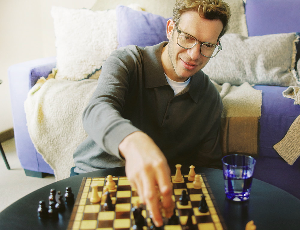 Man met bril speelt schaak in een woonkamer, ontspannen sfeer met paarse kussens op de achtergrond.