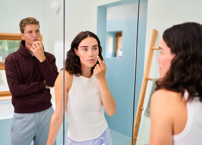 onge vrouw doet contactlens in voor de spiegel, terwijl een jonge man toekijkt in de badkamer.
