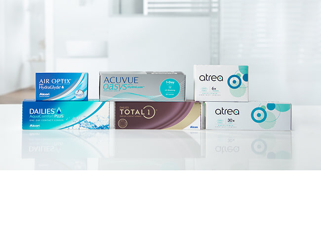 Des emballages de lentilles journalières et mensuelles sur la table, avec une salle de bain visible en arrière-plan.