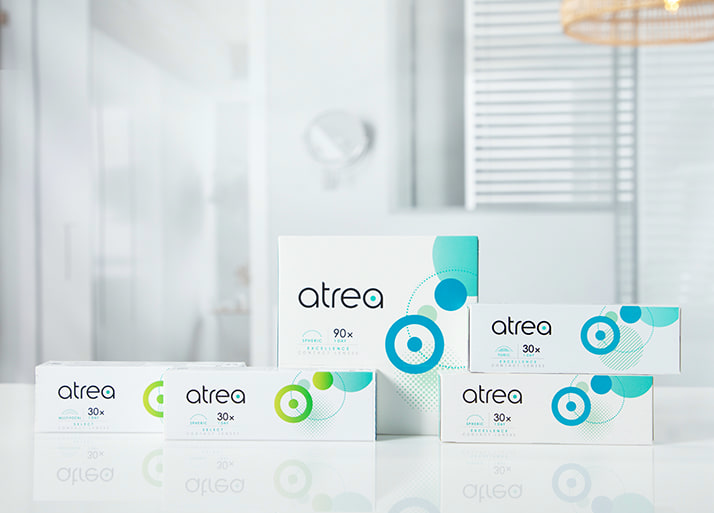 Atrea-contactlenzen verpakkingen in verschillende groottes en typen op een witte ondergrond, met een badkamer op de achtergrond.