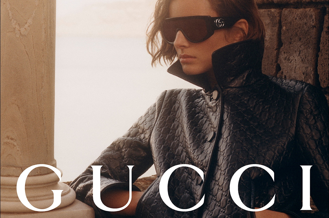 Mannequin en manteau Gucci noir et lunettes logo, ambiance luxueuse face à la mer.