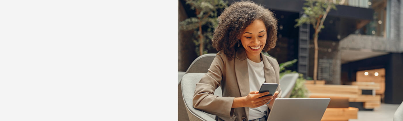 Vrouw met krullend haar zit met een laptop op schoot en kijkt glimlachend naar haar smartphone in een moderne ruimte.