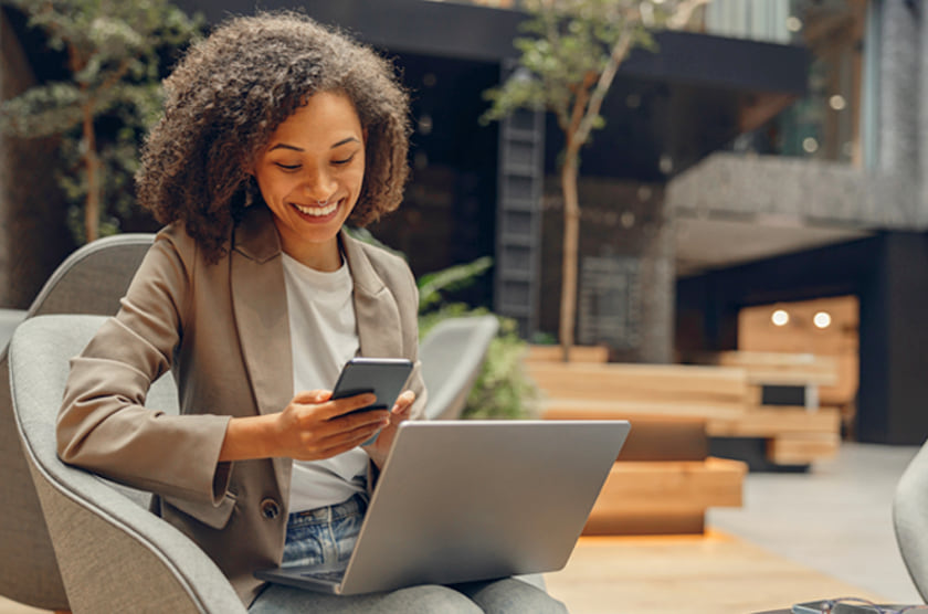 Vrouw met krullend haar zit met een laptop op schoot en kijkt glimlachend naar haar smartphone in een moderne ruimte.