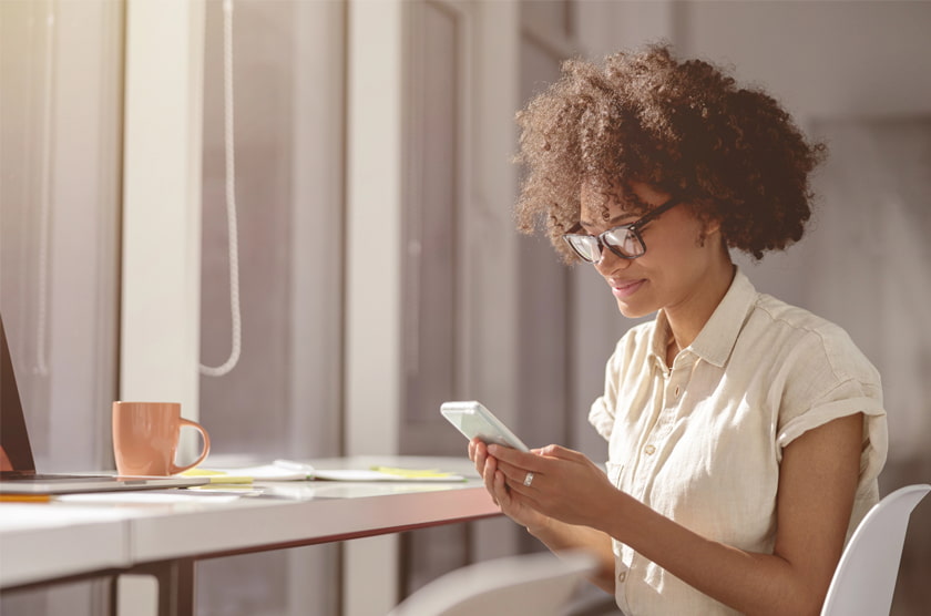 Vrouw met bril zit aan een bureau en kijkt op haar smartphone bij daglicht.