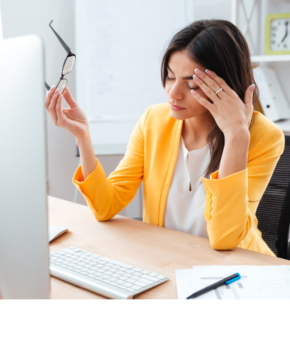 Een vrouw in een gele blazer zit aan een bureau en wrijft vermoeid in haar ogen terwijl ze haar bril in de hand houdt. Voor haar staan een computerscherm, een toetsenbord en kantoorbenodigdheden.