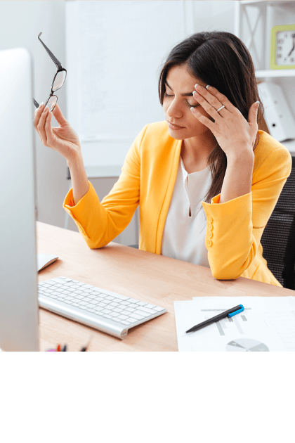 Een vrouw in een gele blazer zit aan een bureau en wrijft vermoeid in haar ogen terwijl ze haar bril in de hand houdt. Voor haar staan een computerscherm, een toetsenbord en kantoorbenodigdheden.