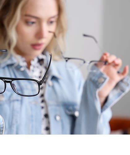 Een jonge vrouw met blond haar en een spijkerjack houdt twee verschillende brillen in haar handen en kijkt er verbaasd naar. Ze lijkt te twijfelen over welke bril ze moet kiezen.