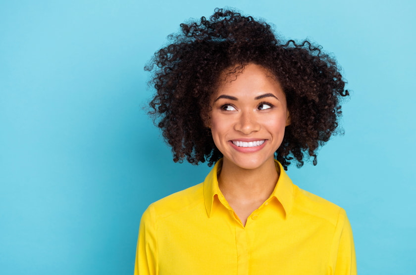 Vrouw met krullend haar en een geel hemd glimlacht en kijkt opzij voor een blauwe achtergrond.