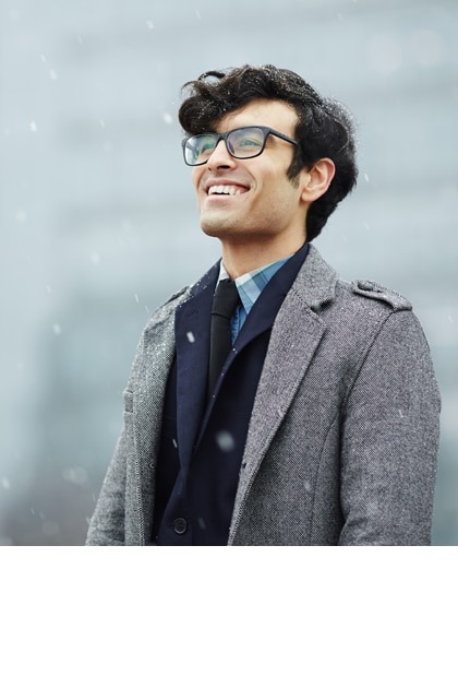 Een jonge man met donker haar en een bril glimlacht en kijkt in de verte terwijl de sneeuw valt. Hij draagt een grijze mantel over een donker pak, met een vervaagde stadsachtergrond.