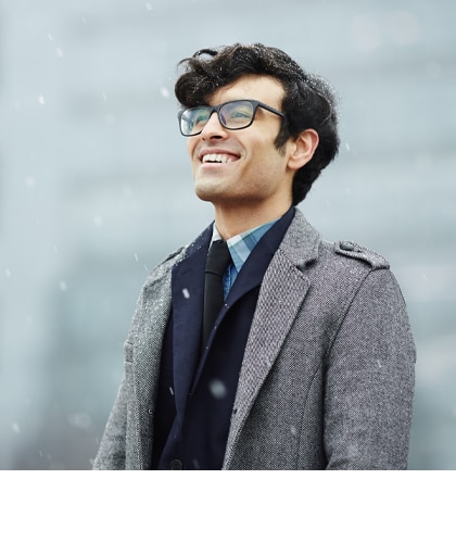 Een jonge man met donker haar en een bril glimlacht en kijkt in de verte terwijl de sneeuw valt. Hij draagt een grijze mantel over een donker pak, met een vervaagde stadsachtergrond.