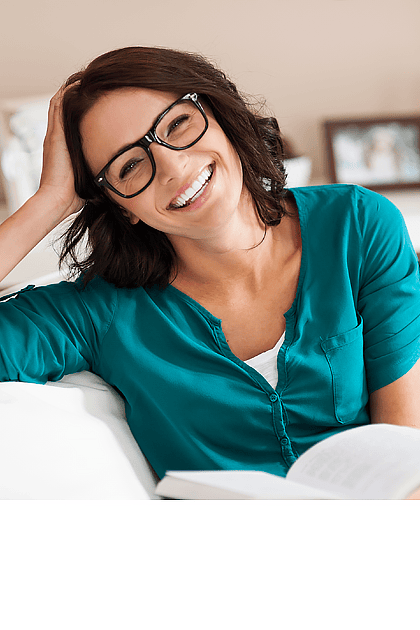 Een lachende vrouw met bruin haar en een zwarte bril leunt ontspannen op een witte bank. Ze draagt een turquoise blouse, terwijl op de achtergrond ingelijste foto\'s en decoratie te zien zijn.