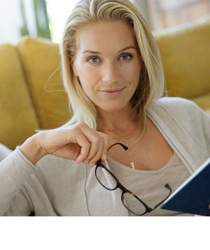 Niederländisch: Een blonde vrouw zit ontspannen op een bank met een boek in de hand en houdt haar bril vast. Ze kijkt rustig en vriendelijk in de camera, met een geel kussen op de achtergrond.