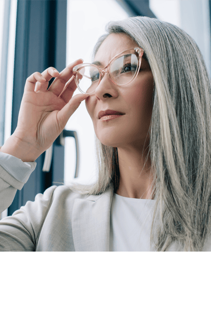 Een vrouw met lang grijs haar en een bril kijkt bedachtzaam uit het raam.