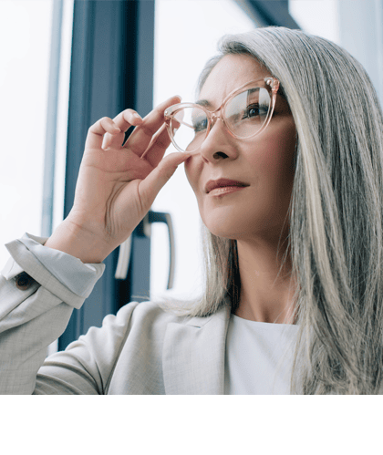 Een vrouw met lang grijs haar en een bril kijkt bedachtzaam uit het raam.