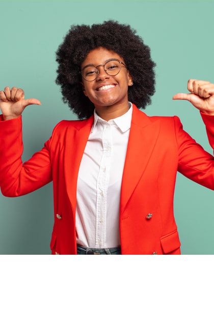 Een vrolijk lachende vrouw met krullend donker haar en een bril wijst met beide duimen naar zichzelf. Ze draagt een rode blazer over een witte blouse en staat voor een mintgroene achtergrond.
