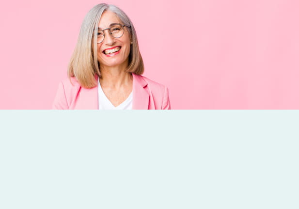 Een oudere vrouw met grijs, schouderlang haar draagt een stijlvolle bril met een dun montuur. Ze lacht vriendelijk en draagt een roze blazer voor een roze achtergrond.