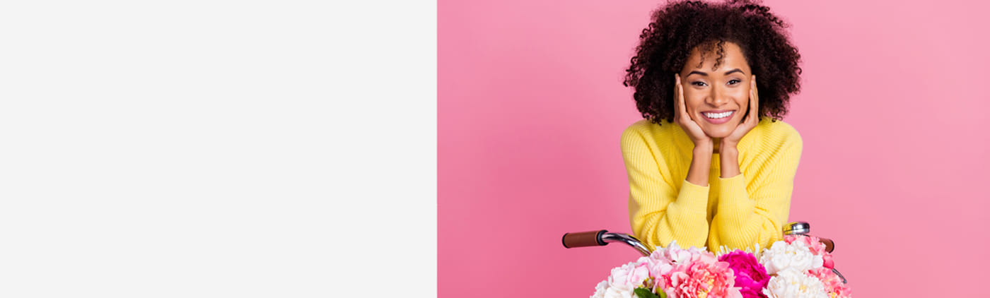 Vrouw met krullend haar en een gele trui glimlacht achter een fiets met bloemen op een roze achtergrond.