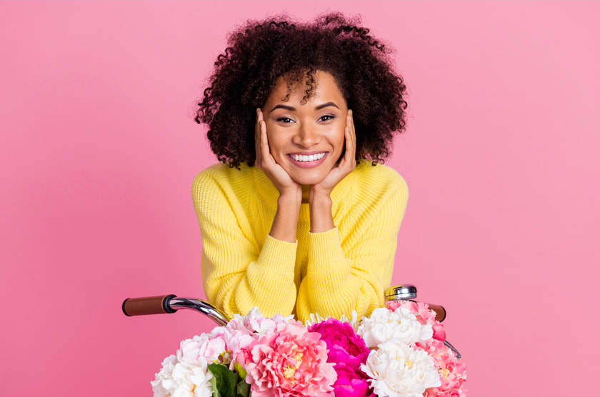 Vrouw met krullend haar en een gele trui glimlacht achter een fiets met bloemen op een roze achtergrond.