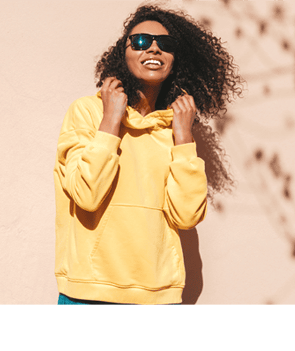 Een lachende vrouw met krullend donker haar draagt een zwarte zonnebril en een gele hoodie. Ze staat voor een beige muur met schaduwen van bladeren.
