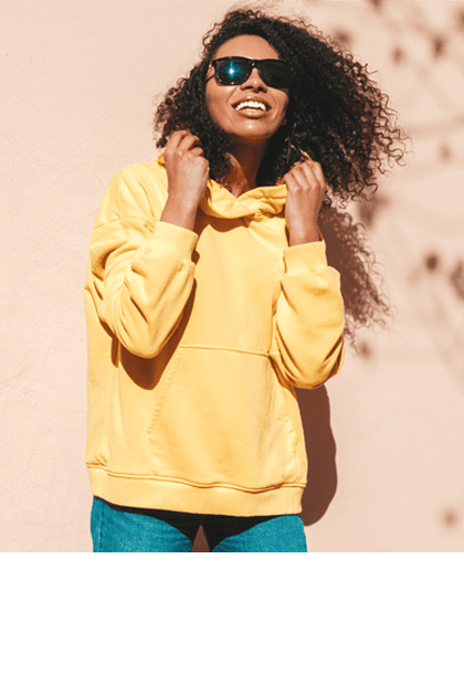 Een lachende vrouw met krullend donker haar draagt een zwarte zonnebril en een gele hoodie. Ze staat voor een beige muur met schaduwen van bladeren.